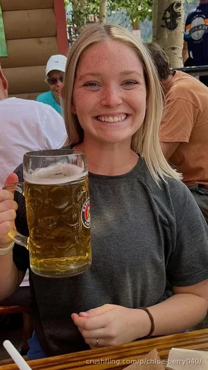 Blonde woman in gray t-shirt holding beer mug, smiling at outdoor seating area.