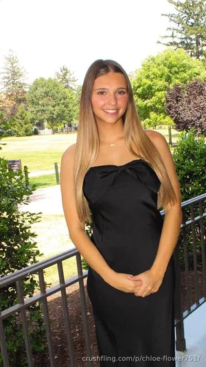 Woman with long blonde hair in black strapless dress standing outdoors near railing and greenery.