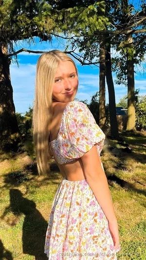 Blonde woman in floral dress outdoors under trees, back turned slightly.