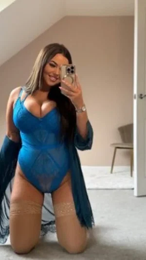 Woman in blue sheer bodysuit and lace garter stockings kneeling indoors for mirror selfie.