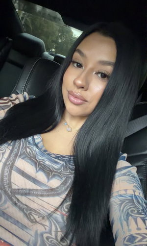 Woman with long black hair in patterned top sitting in car.