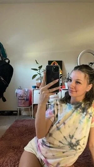 Woman in tie-dye shirt and shorts taking mirror selfie in bedroom.