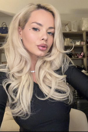 Woman with long blonde hair in black top, posing indoors in front of shelves.