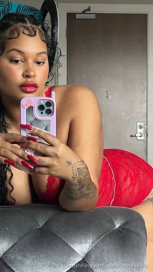 Woman in red lace lingerie taking mirror selfie in bedroom, curly hair, tattooed arm.
