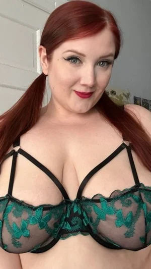 Woman with red hair in a green lace bra, indoors, facing camera.