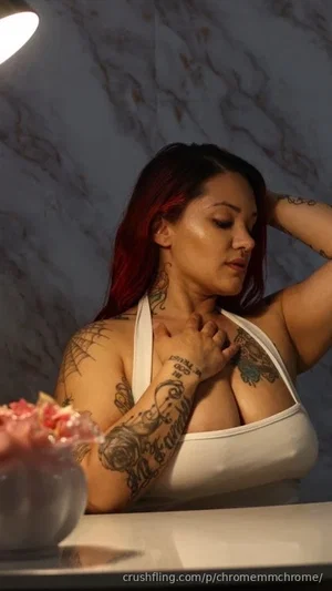 Woman with red hair and tattoos wears white top, posing indoors near marble wall.