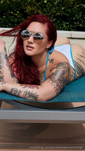 Woman with red hair and tattoos in a blue bikini top, lounging outdoors on a chair.