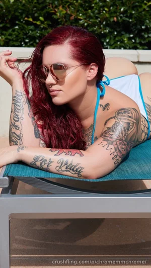 Woman with red hair and tattoos in a blue bikini top lounging outdoors on a chair.