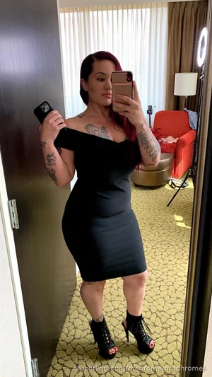 Woman with tattoos in black off-shoulder dress and heels, taking mirror selfie in hotel room.
