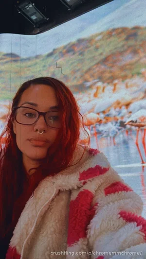 Woman with red hair and glasses wearing a pink and white fuzzy jacket, standing in front of a