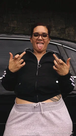 Woman with glasses and tattoos sticks tongue out, wearing black jacket and gray sweatpants outdoors.