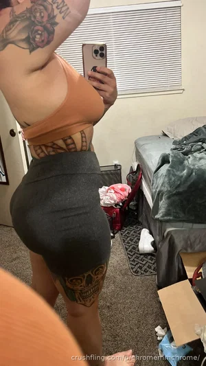 Woman with tattoos in orange top and gray shorts taking mirror selfie in bedroom.