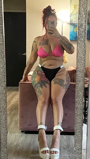 Woman with tattoos in pink bikini top and black bottoms, taking mirror selfie in bedroom.