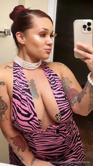 Woman with tattoos in pink zebra-print dress, pearls, mirror selfie, indoor setting.