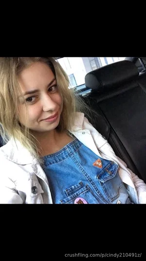 Woman with blonde hair wearing denim overalls and white jacket, sitting in a car.