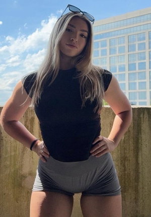 Blonde woman in black top and gray shorts posing outdoors.