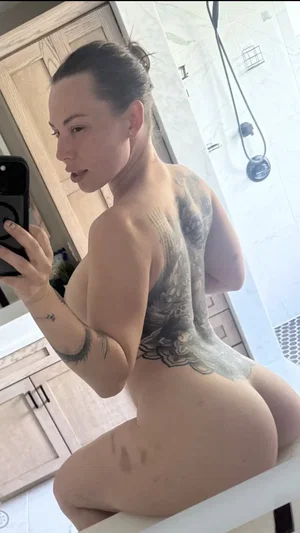 Woman with tattoos poses nude in bathroom, back to camera, holding phone for mirror selfie.