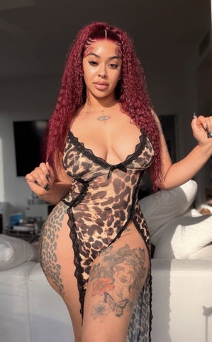 Woman with red curly hair in leopard print sheer bodysuit, standing indoors near white couch.