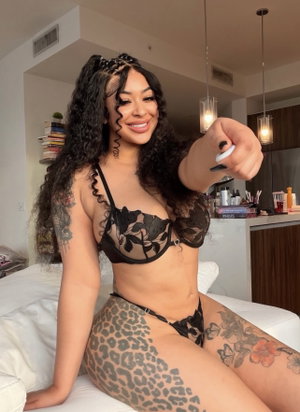 Woman with curly hair and tattoos wears black lace lingerie, sitting on a white couch indoors.