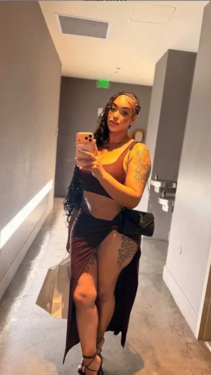Woman with tattoos in a brown crop top and high-slit skirt taking a mirror selfie in a hallway.