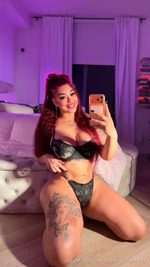 Woman with red hair in black lace lingerie posing on floor in bedroom with purple lighting.