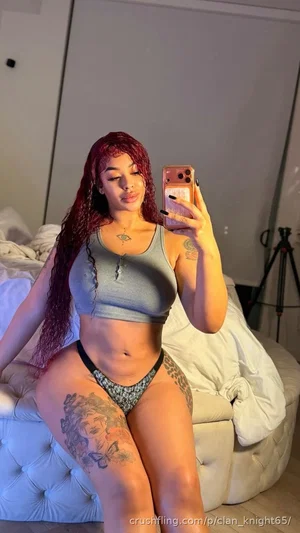 Woman with red hair and tattoos wears gray crop top and patterned underwear, sitting on bed in