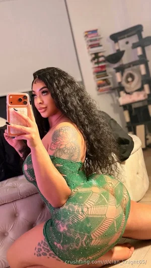 Woman with long dark hair in green sheer dress, sitting on floor, mirror selfie in bedroom.