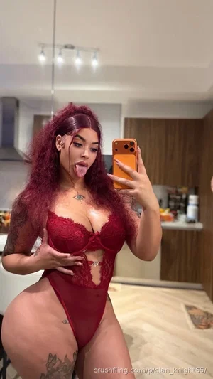 Woman with red hair in red lace lingerie posing in kitchen, tongue out, tattoos visible.