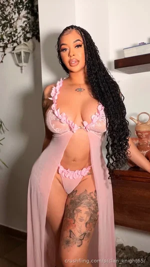 Woman with long black hair in pink lace lingerie and sheer robe, standing indoors with tattoos