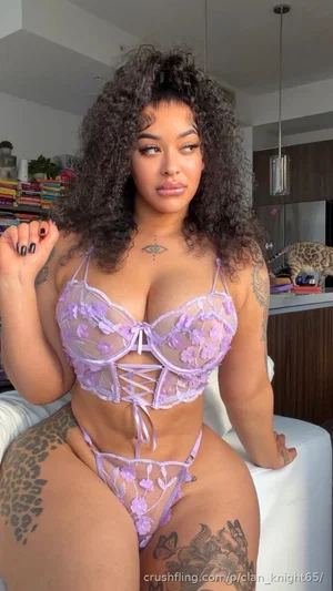 Woman with curly hair in purple lace lingerie, sitting indoors with tattoos visible.