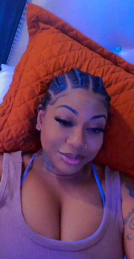 Woman with braided hair and tattoos wearing a tank top, lying on an orange pillow indoors.