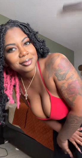 Woman with curly hair and tattoos wears red top, posing indoors near bed.