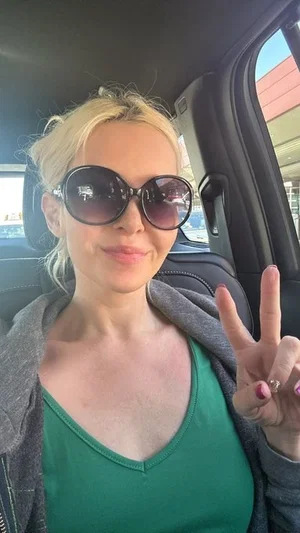 Blonde woman in green top and gray hoodie, wearing sunglasses, making peace sign in car.
