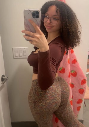 Woman with curly hair in brown top and patterned leggings, posing in mirror selfie indoors.