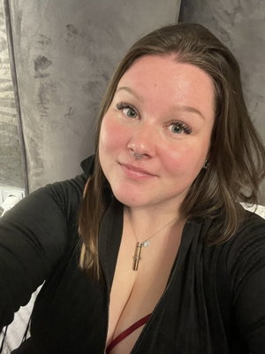 Woman with brown hair and nose ring wearing black top, taking selfie indoors.