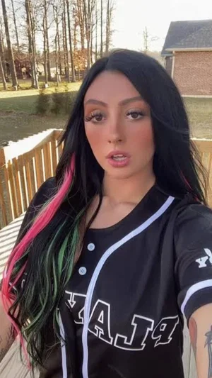 Woman with long black hair and pink green streaks wears black baseball jersey, outdoors on wooden