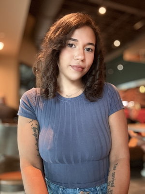 Woman with curly hair in blue ribbed top and jeans, standing indoors with tattoos on arms.