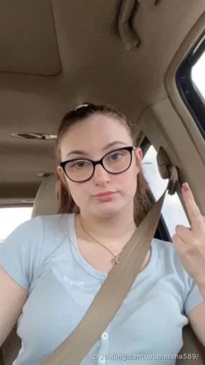 Woman with glasses and nose ring in car, wearing light blue shirt, giving middle finger.
