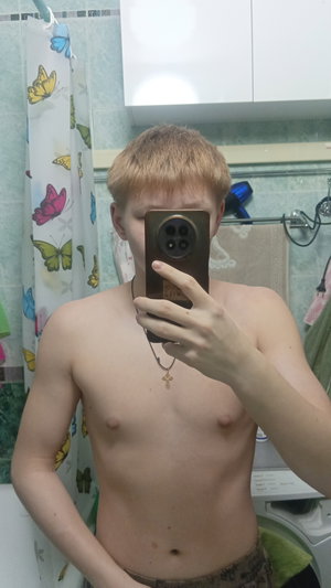 Slim blond man shirtless in camo pants bathroom selfie.