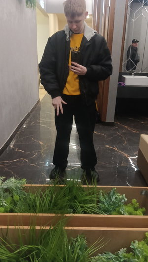 Man in black jacket, yellow hoodie, black pants in bathroom.