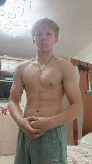 Shirtless man with defined muscles wearing green pants, standing indoors in a bedroom.
