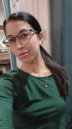 Dark-haired woman with glasses in green top taking selfie indoors.