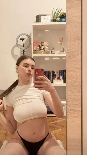 Woman in beige crop top and black underwear taking mirror selfie in bedroom.