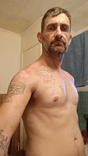 Shirtless man with tattoos on chest and arm, standing indoors in a bathroom.