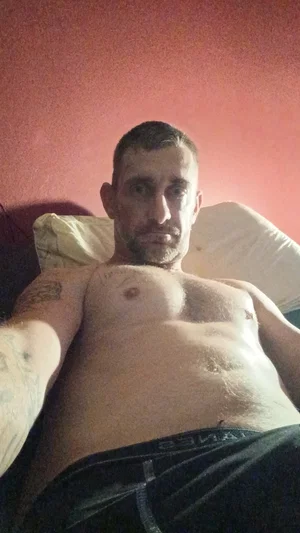 Shirtless man with tattoos lying on bed, wearing dark pants, red wall behind.
