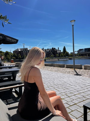 Blonde woman in black dress sitting outdoors by the water.