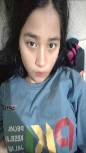 Asian woman with long dark hair in blue t-shirt taking indoor selfie.