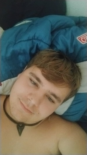 Shirtless man with short brown hair lying on bed, wearing black choker, blue comforter in