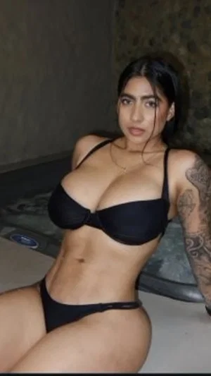 Woman in black lingerie posing indoors, tattooed arm visible.