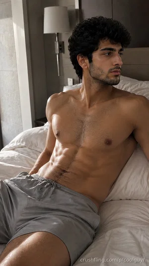 Shirtless man with curly hair and defined abs sitting on a bed in gray shorts.